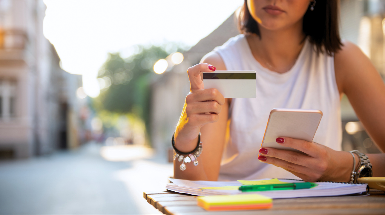 Woman holding credit card and phone outdoors while shopping online